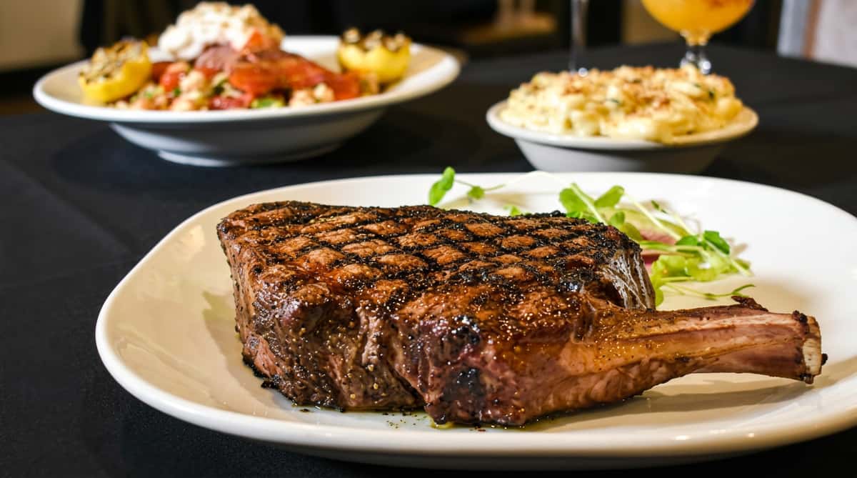 Grilled ribeye steak with grill marks and microgreens on a white plate, with sides and drink blurred in background.