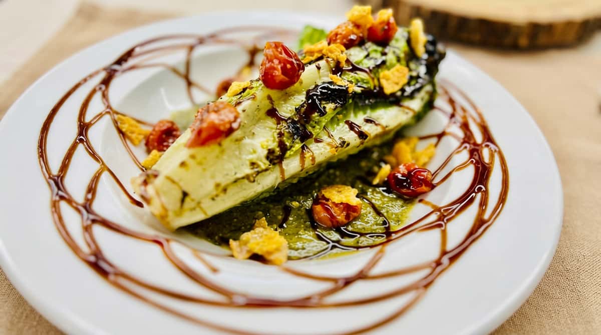 Grilled romaine heart with cherry tomatoes, croutons, green pesto, and balsamic glaze on a white plate.