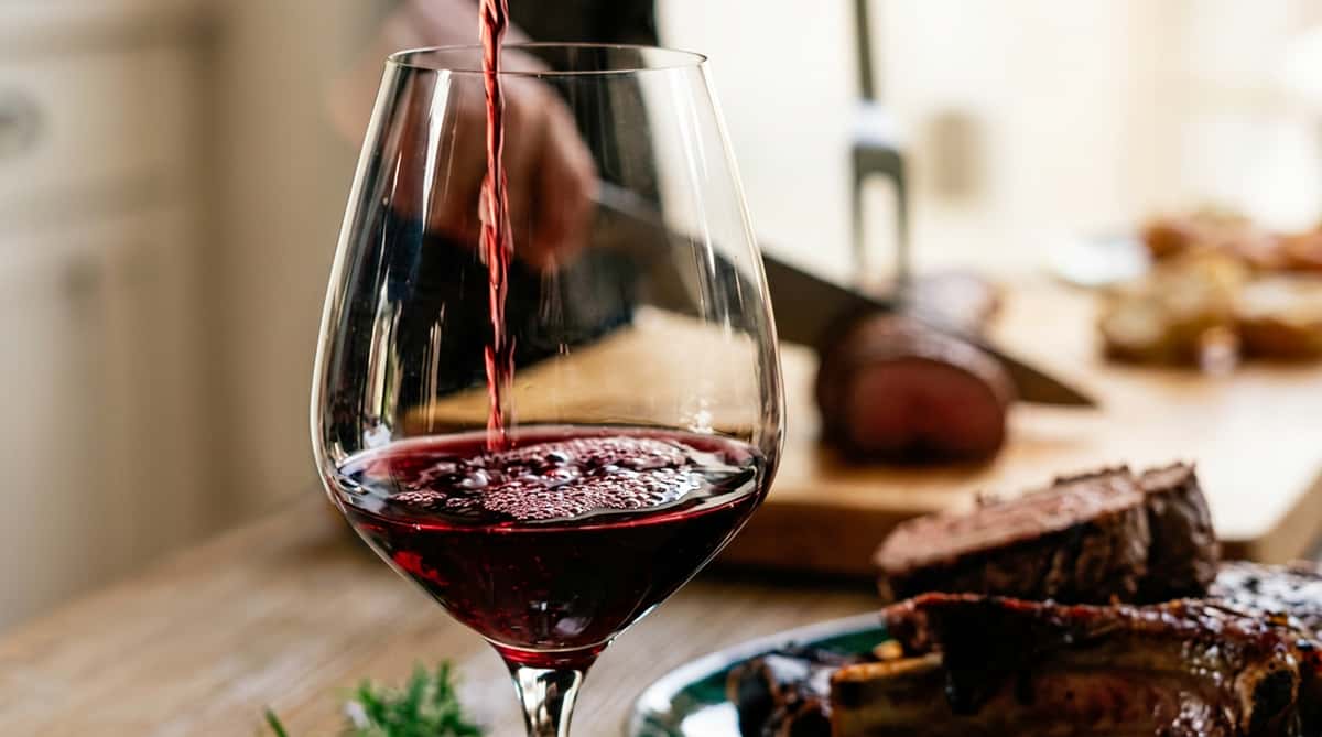Red wine being poured into a clear glass, with a blurred background showing sliced meat and a carving knife.