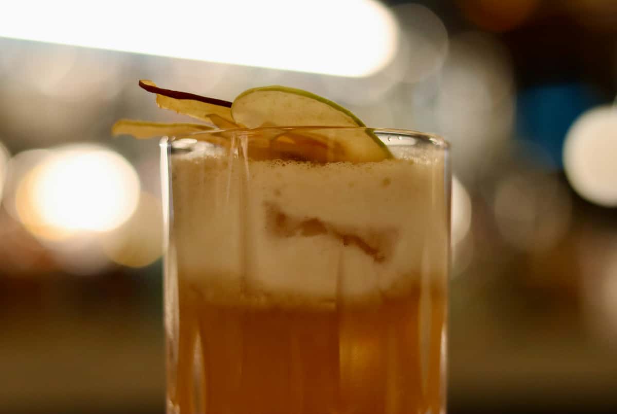 Foamy cocktail in a tall glass garnished with thin apple slices, set against a softly blurred background of warm ambient lights.