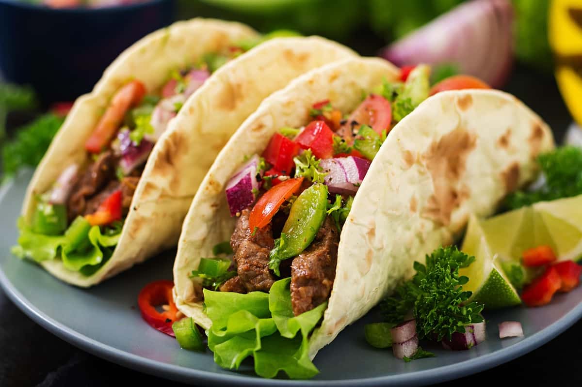 Three beef tacos with lettuce, diced red onion, tomatoes, and avocado slices on soft flour tortillas with lime wedges on the side.