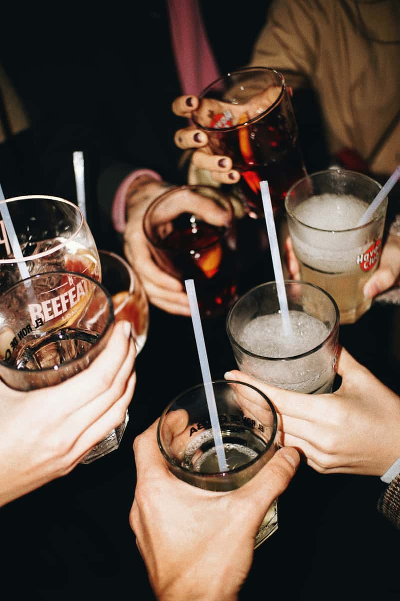 Group of friends raising mixed drinks with straws in a celebratory toast, captured from above in a lively, dimly lit setting.