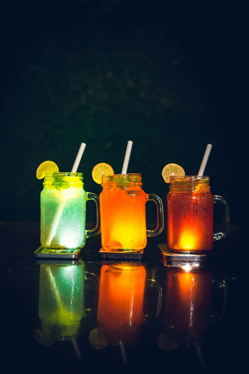 Three colorful drinks in mason jars glow green, orange, and red, each garnished with a lemon slice and served with a straw.