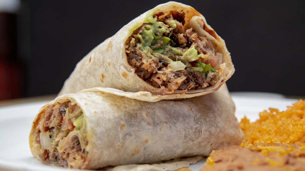 A hearty burrito filled with shredded beef, guacamole, and fresh vegetables wrapped in a soft flour tortilla, served with a side of rice and refried beans.