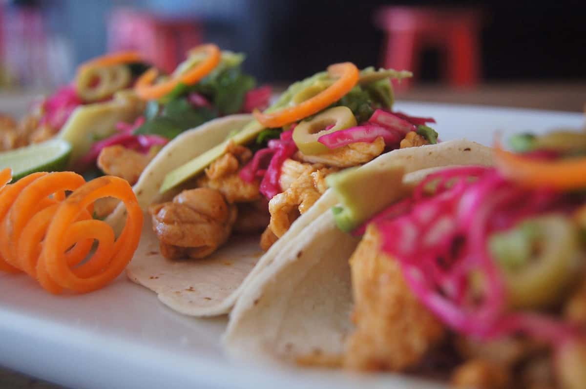 Fresh chicken tacos topped with pickled red onions, avocado slices, green olives, and garnished with curly carrot ribbons on a white plate.