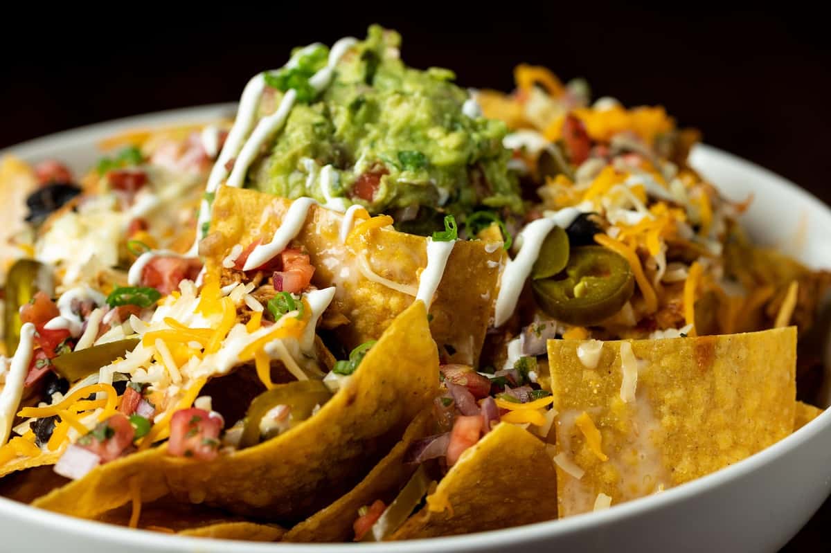 A loaded plate of nachos topped with shredded cheese, jalapeños, pico de gallo, sour cream drizzle, and a generous scoop of guacamole.