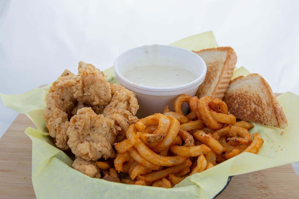 Basket of fried chicken tenders, seasoned curly fries, toasted bread slices, and a cup of ranch dipping sauce on yellow paper.