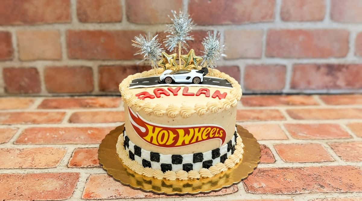 Hot Wheels cake with a white toy car, silver sparklers, and golden stars on top, "RAYLAN" and logo on the side.