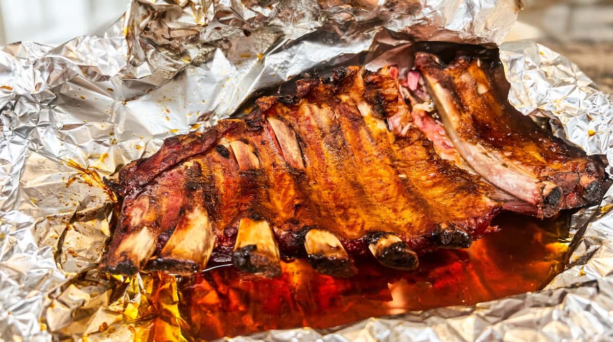 Rack of cooked ribs with a reddish-brown crust, resting in a pool of red liquid inside foil.