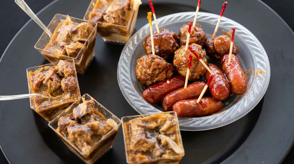 A clear plastic plate of glazed meatballs and cocktail sausages with toothpicks, next to small square cups of a brown dessert.