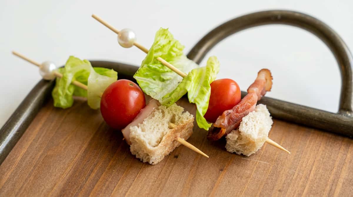 Appetizer skewers with cherry tomatoes, lettuce, slivers of prosciutto, and bread cubes on a wooden board.