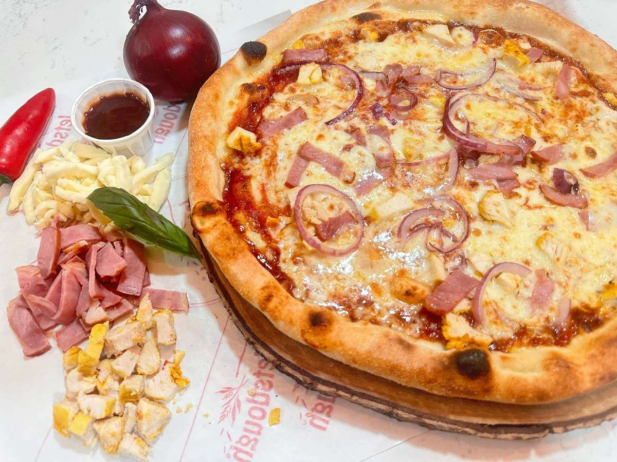 Stone-baked pizza with ham, red onions, melted cheese, and barbecue-style sauce, served with fresh ingredients on the side