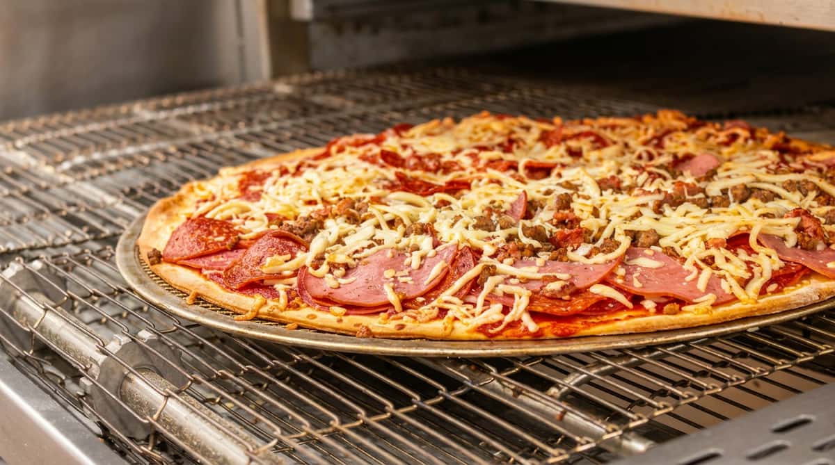 Uncooked pizza with pepperoni, ham, crumbled sausage, and shredded mozzarella cheese in an oven.