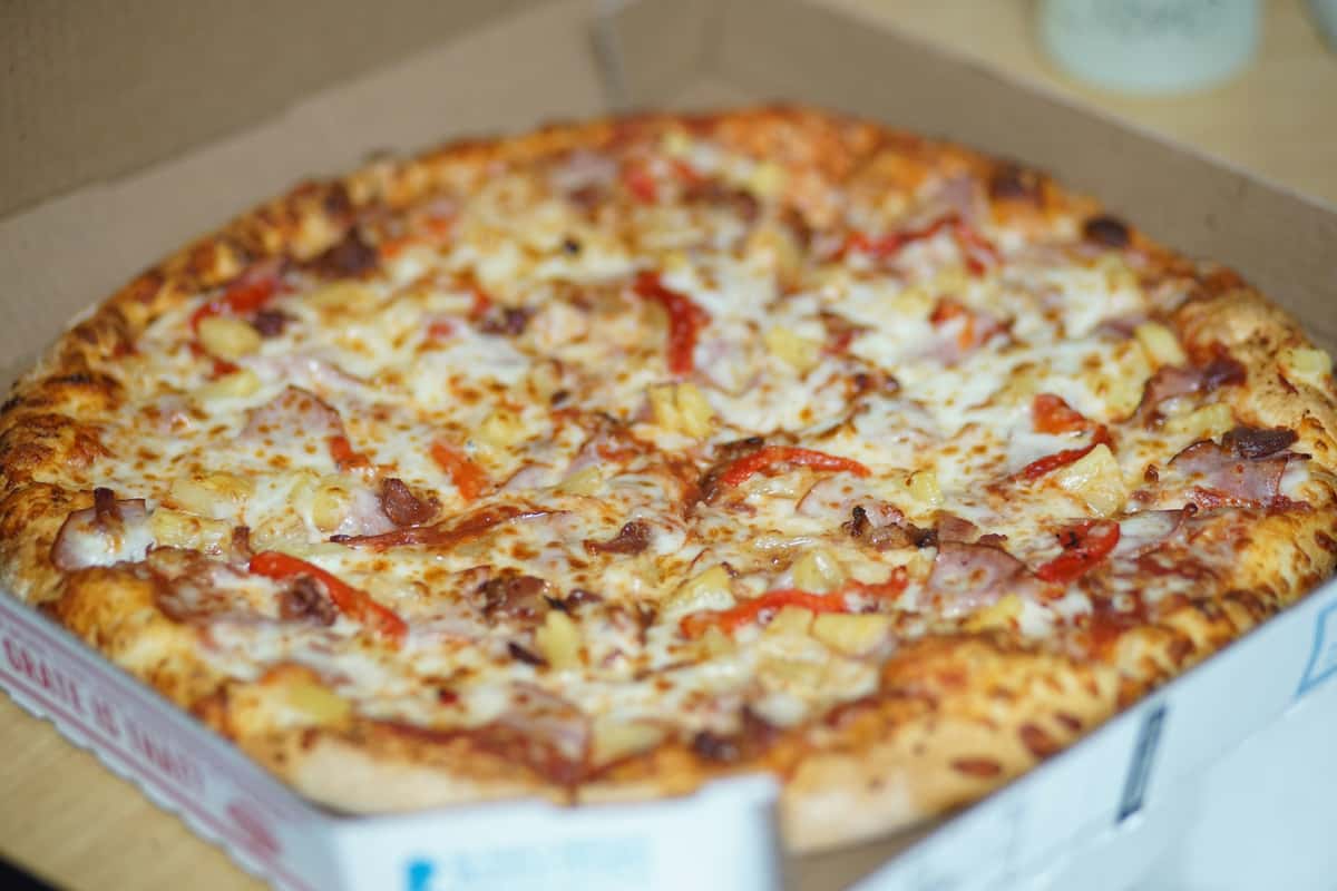 Freshly baked Hawaiian pizza in an open delivery box, topped with ham, pineapple, red bell peppers, and melted mozzarella cheese.