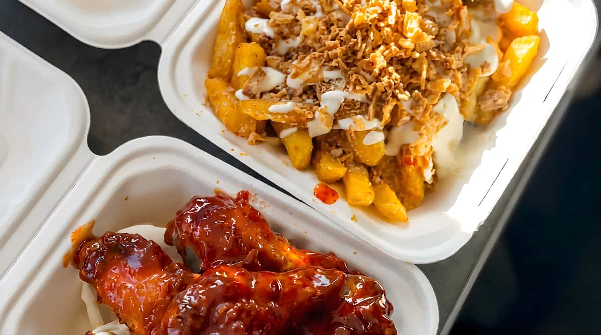Takeout container with sauced chicken wings and loaded fries topped with cheese and crispy onions
