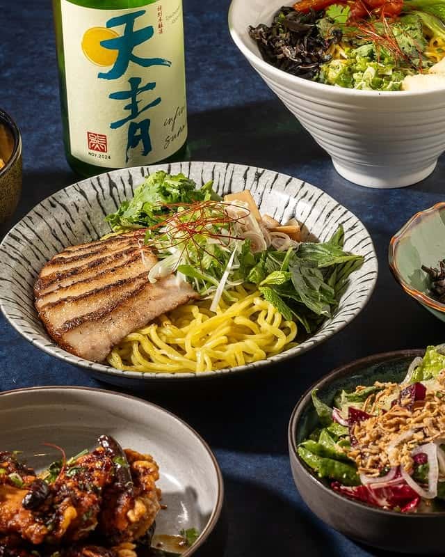 Japanese ramen spread featuring grilled pork over yellow noodles with fresh herbs and bamboo shoots, surrounded by small plates and salads, with a bottle of sake for a modern Japanese restaurant dining experience.