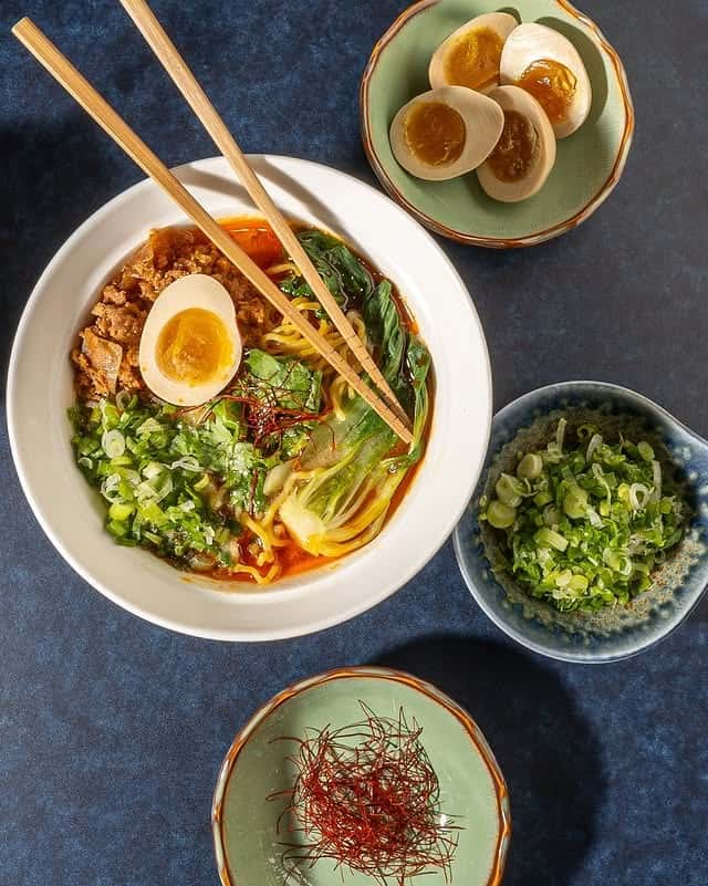 Spicy Japanese ramen bowl with chili broth, wheat noodles, minced pork, bok choy, scallions, and a halved soy-marinated egg, photographed overhead with chopsticks and small side dishes for a modern Asian restaurant presentation.