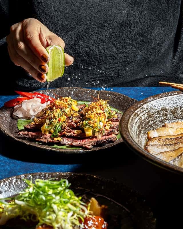 Hand squeezing fresh lime over sliced seared beef topped with chili, herbs, and sauce, plated on a dark ceramic dish with Japanese-inspired sides, captured in dramatic lighting for an upscale Asian restaurant dining scene.