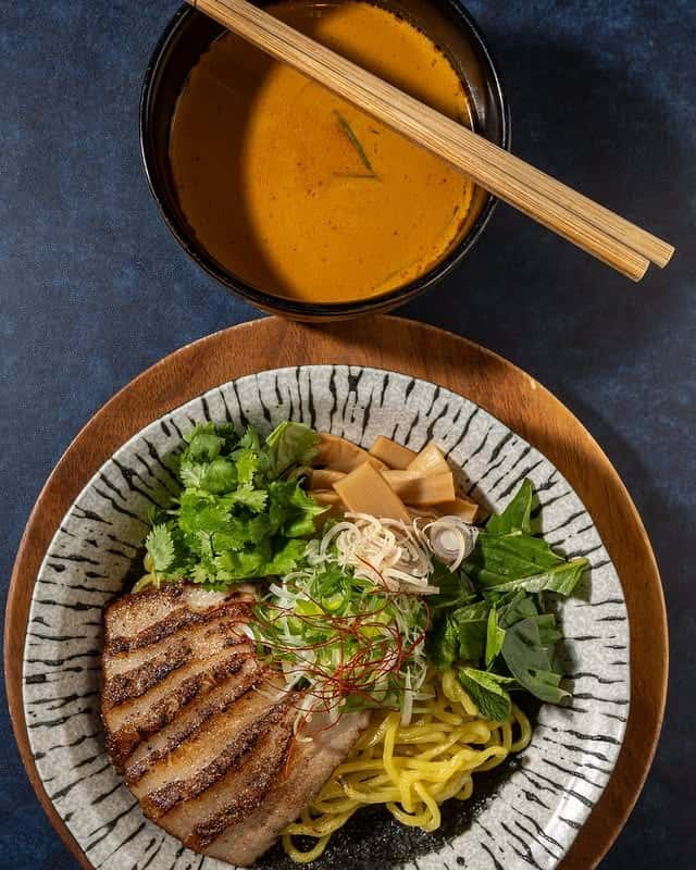 Japanese dipping ramen set with grilled pork belly slices, yellow wheat noodles, fresh cilantro, scallions, bamboo shoots, and leafy greens, served with a rich orange dipping broth and chopsticks in a modern Asian restaurant setting.