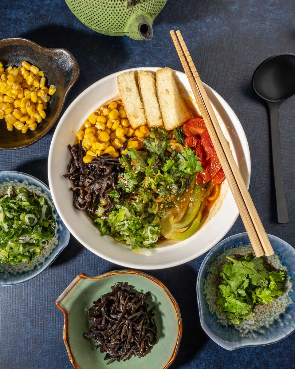 Overhead view of Japanese ramen bowl topped with sweet corn, wood ear mushrooms, scallions, bok choy, tofu strips, and chili oil, styled with chopsticks, teapot, and small side dishes for a modern Japanese restaurant presentation.