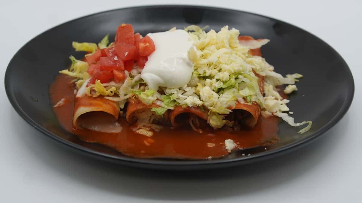 Three rolled enchiladas topped with red enchilada sauce, shredded lettuce, diced tomatoes, sour cream, and cheese—served on a black plate for a classic Mexican comfort dish.