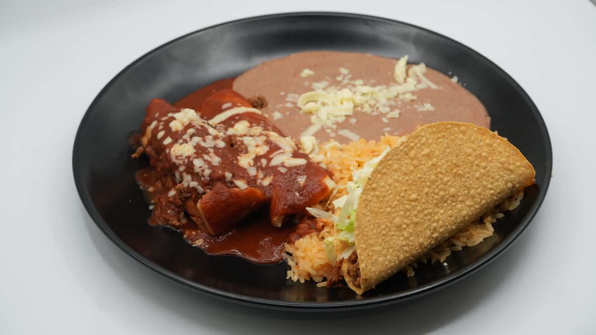 Mexican combination plate featuring a beef enchilada smothered in red sauce and melted cheese, a crunchy ground beef taco with lettuce, Mexican rice, and refried beans topped with shredded cheese—served on a black dish for a hearty, traditional meal.
