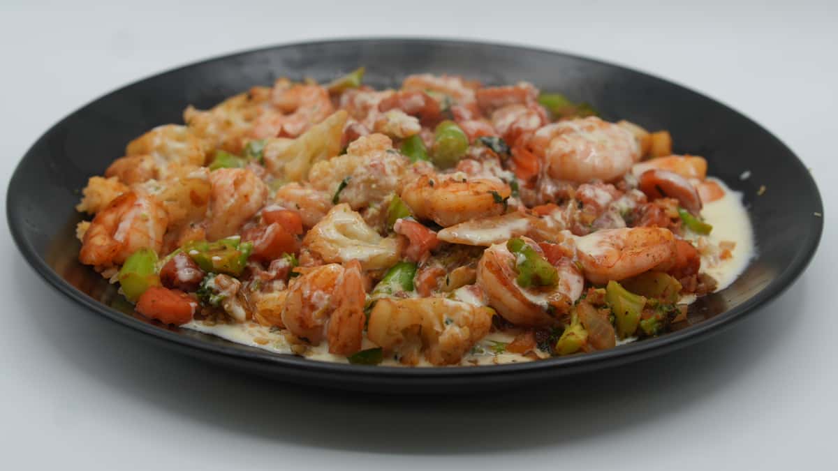 Sautéed shrimp, broccoli, cauliflower, bell peppers, and onions smothered in creamy queso sauce—served sizzling on a black plate for a flavorful seafood and veggie medley.