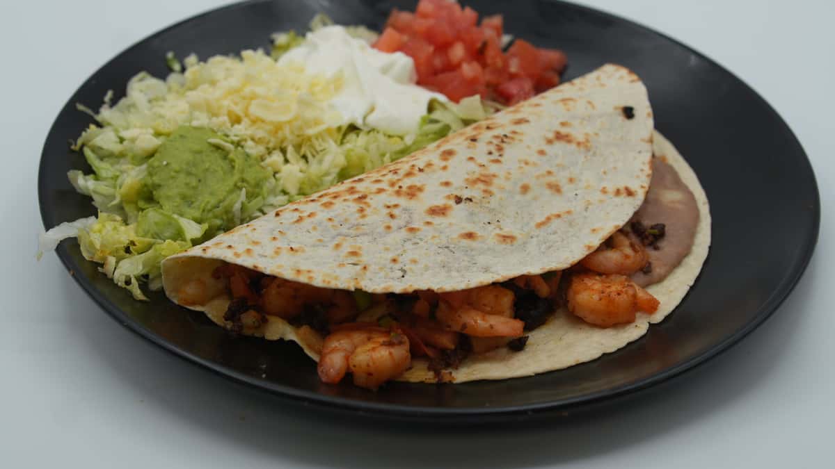 Grilled shrimp taco served on a lightly toasted flour tortilla with refried beans, accompanied by shredded lettuce, guacamole, sour cream, shredded cheese, and diced tomatoes—presented on a black plate for a flavorful and colorful Mexican dish.