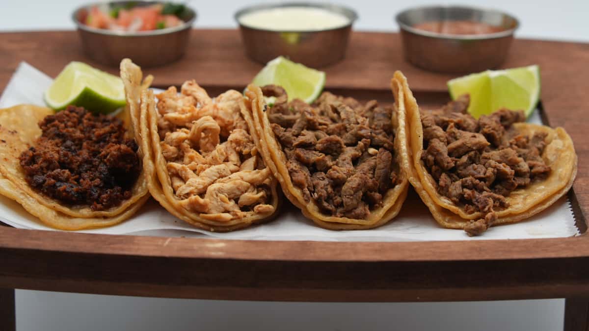 Close-up of a wooden tray featuring four street-style tacos with a variety of meats, including chorizo, grilled chicken, and seasoned beef, served on double corn tortillas with lime wedges. In the background are small bowls of fresh salsa, queso, and pico de gallo, typical of an authentic Mexican grill.