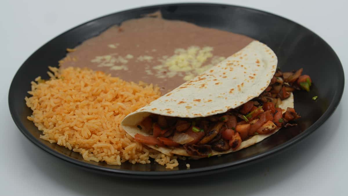 Grilled chicken and vegetable taco folded in a warm flour tortilla, served with a generous portion of Mexican rice and refried beans topped with melted cheese on a black plate—classic comfort food from a traditional Mexican grill.