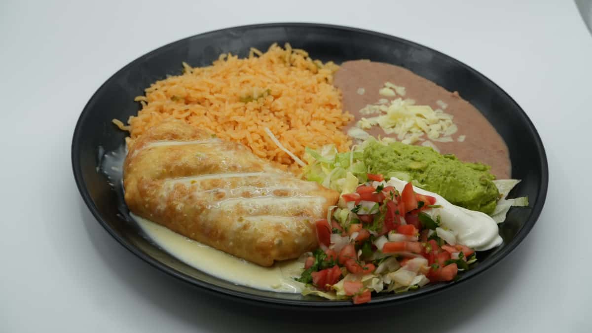 Crispy chimichanga topped with queso sauce, served with Mexican rice, refried beans with shredded cheese, and a fresh side of lettuce, guacamole, sour cream, and pico de gallo—plated on a black dish for a colorful and satisfying Mexican entrée.