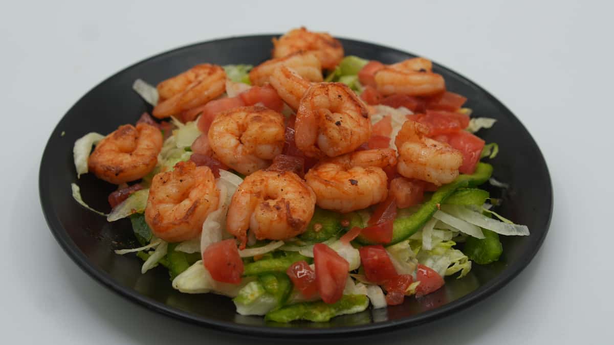 Black plate filled with a vibrant shrimp salad featuring grilled, seasoned shrimp atop a bed of shredded lettuce, sliced green bell peppers, and diced tomatoes—offering a fresh, protein-packed option at a Mexican seafood grill.