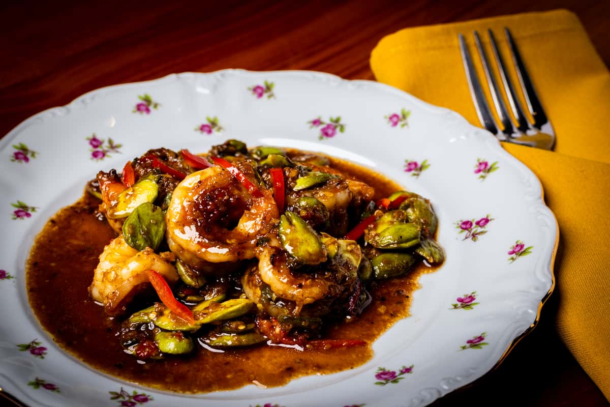 Shrimp sautéed in glossy brown chili sauce with sliced bitter melon and red peppers on a floral white plate beside a yellow napkin.