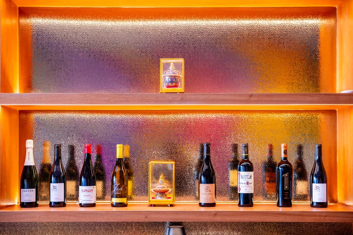 Backlit wooden shelves displaying assorted wine bottles and decorative jars against textured glass with warm amber lighting