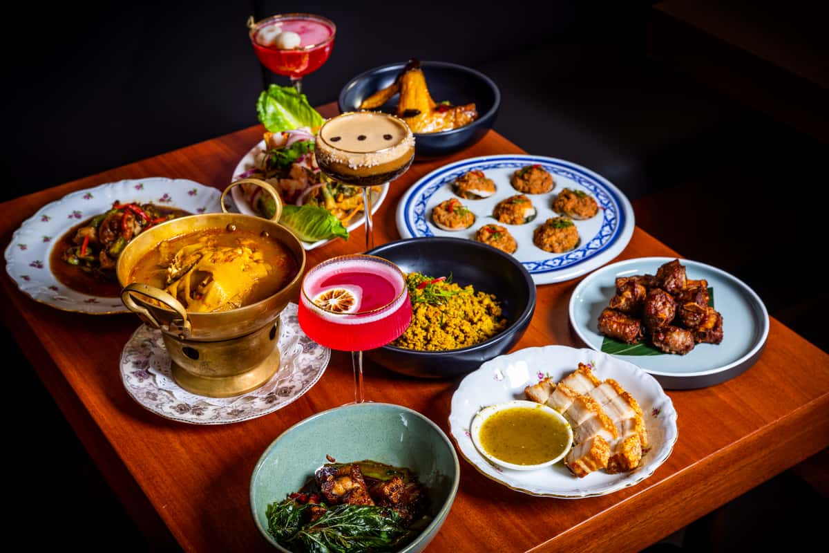 Colorful spread of Thai dishes and craft cocktails arranged on a wooden table, featuring curries, fried bites, rice, and vibrant drinks.