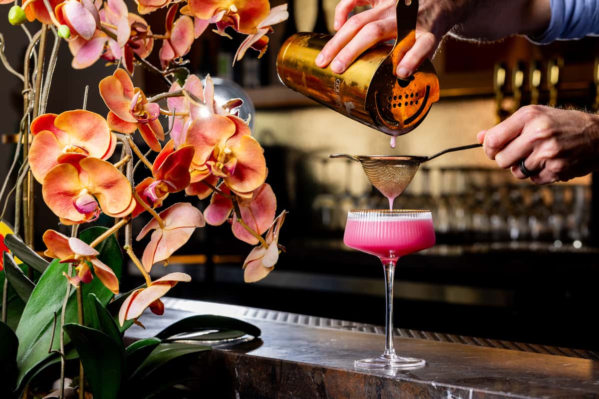 Bartender strains a pink cocktail into a coupe glass beside blooming orchids on a bar counter.
