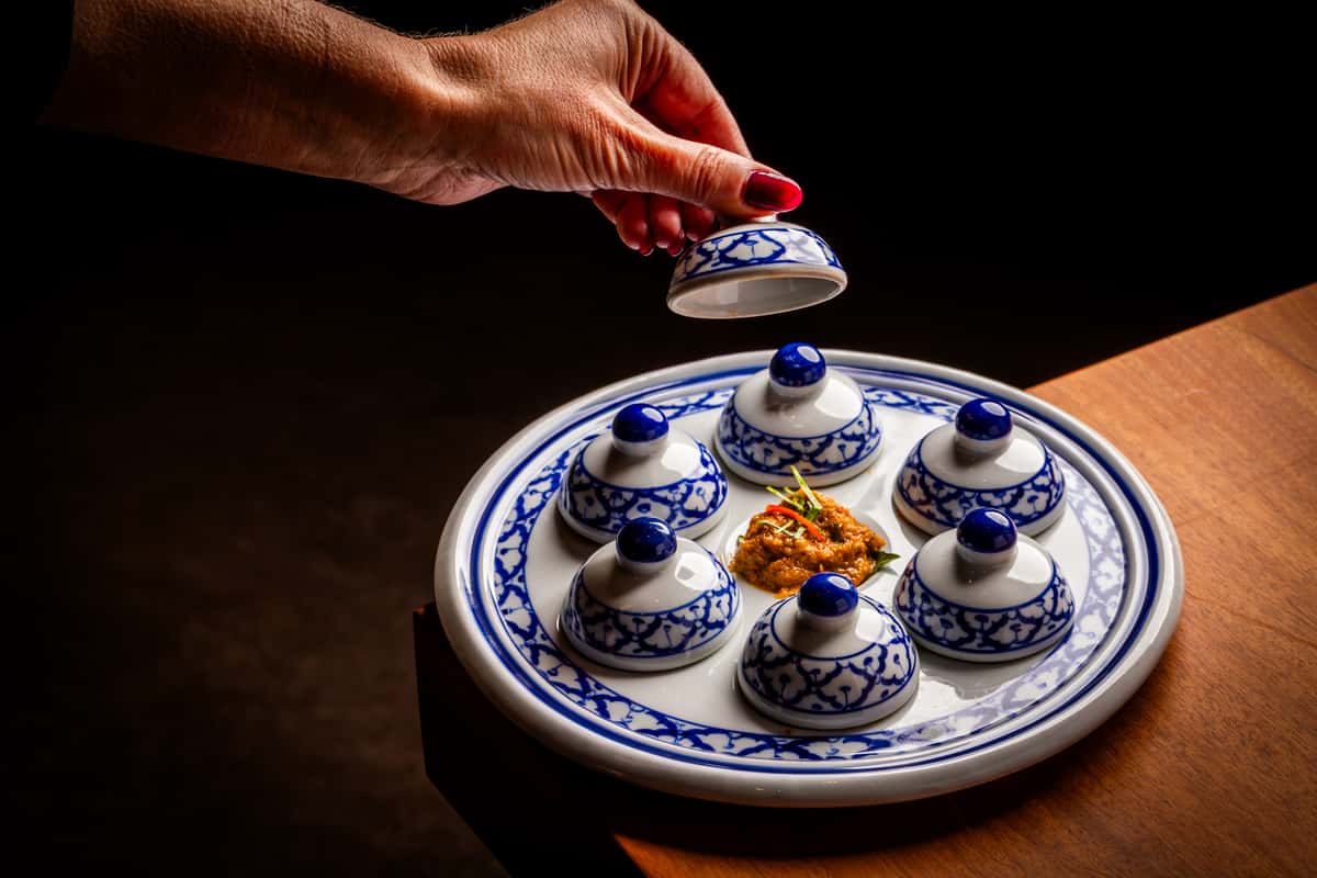 Blue-and-white ceramic platter with six covered dishes arranged around a central dollop of spicy relish on a wooden table.