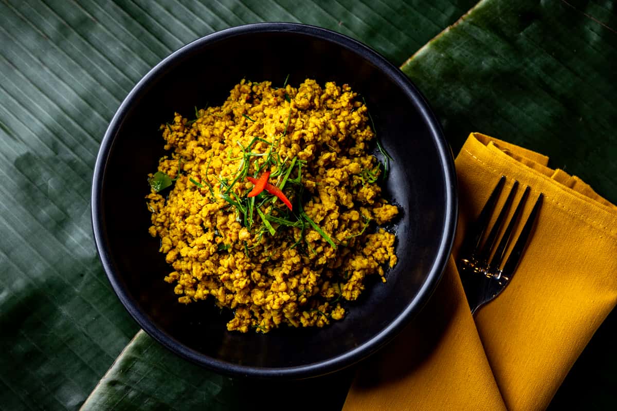 Yellow minced curry garnished with herbs and red chili in a black bowl, set on a textured banana leaf background