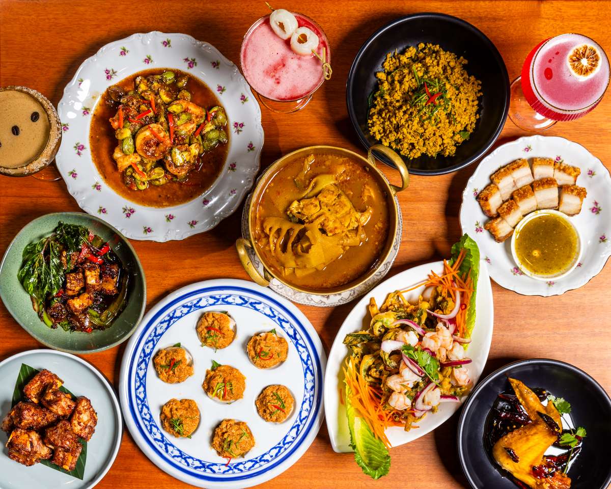 Overhead spread of Thai dishes and colorful cocktails on a wooden table with curries, seafood, pork, and small plates.
