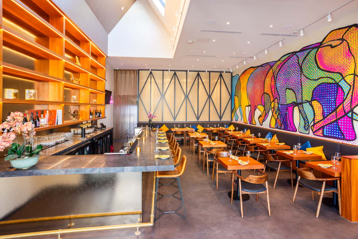 Modern restaurant interior with marble bar, warm wood shelving, set tables, and a colorful elephant mural along the dining wall