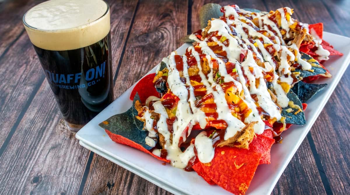 Nachos with red and blue tortilla chips, pulled pork, BBQ sauce, and ranch dressing, next to a pint of dark beer.