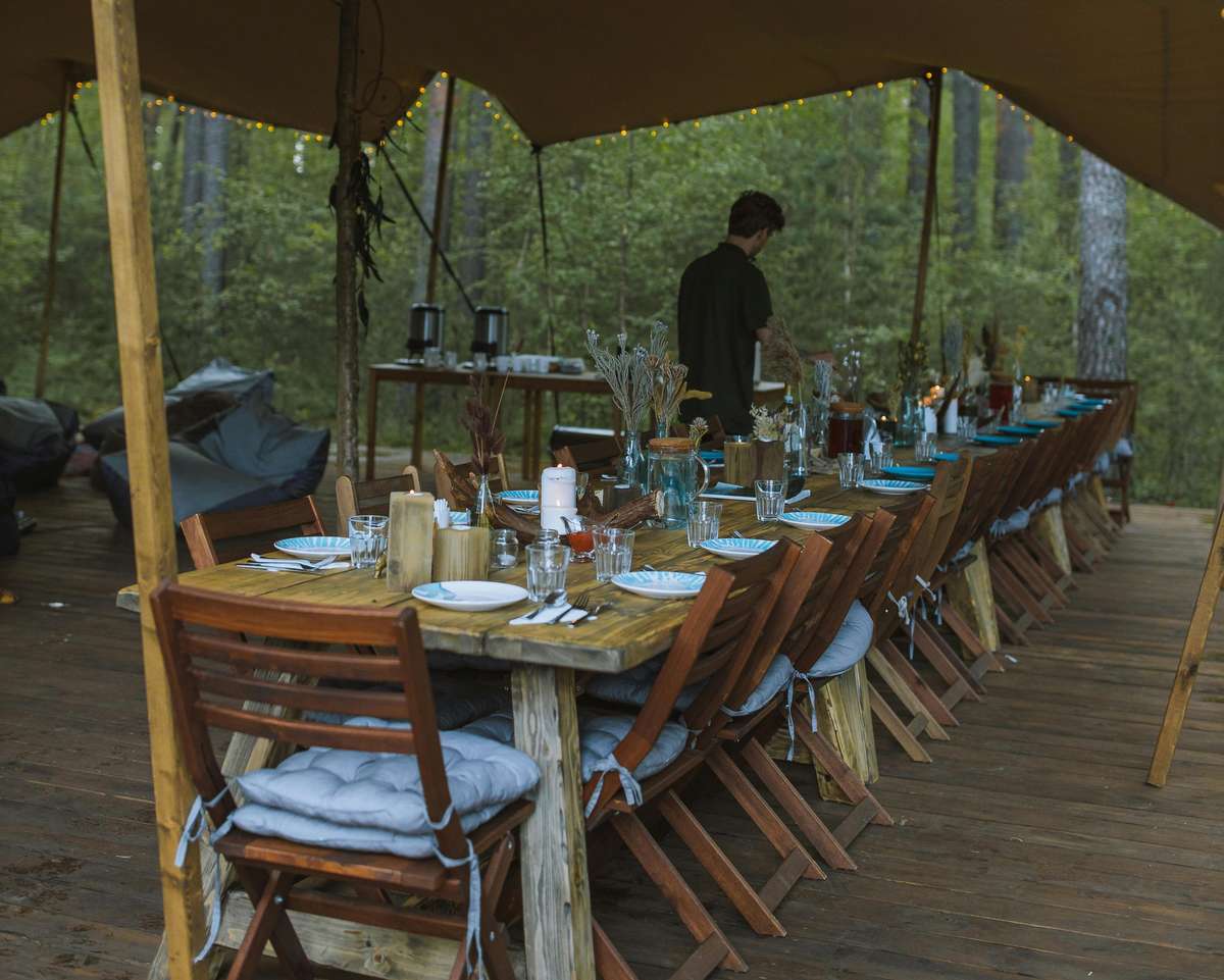 A person prepares the long, set table at Pierce Creek's outdoor dining area, complete with wooden chairs, place settings, and decorative centerpieces.