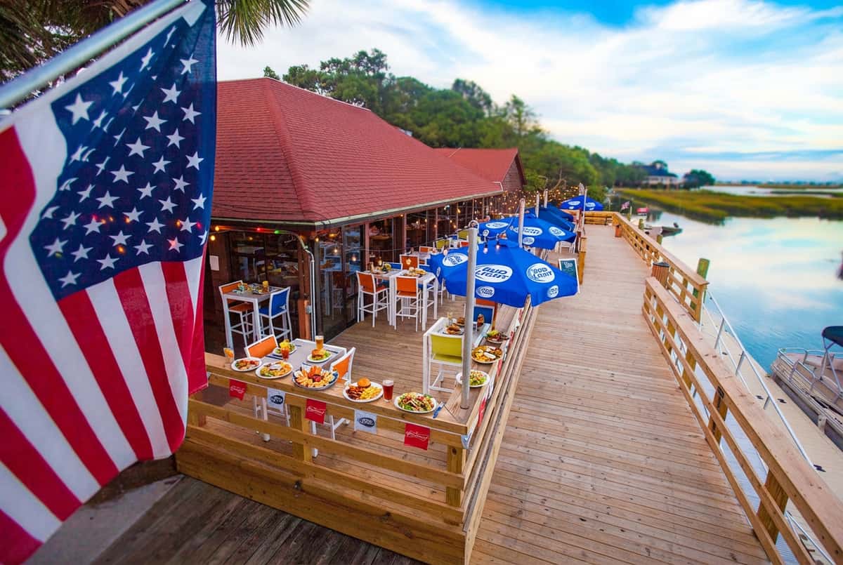 Expansive waterfront dining deck at Bovine's in Murrells Inlet, SC, lined with fresh seafood and wood-fired dishes, offering dockside seating under vibrant blue umbrellas and American flags