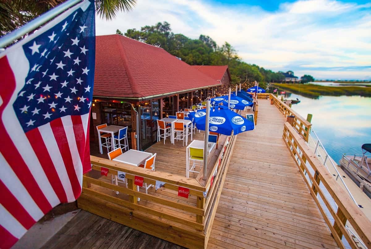 Rustic sunset patio dining setup at Bovine's in Murrells Inlet, SC, with colorful seating, fresh plates of coastal cuisine, and string lights creating a relaxed waterfront atmosphere.