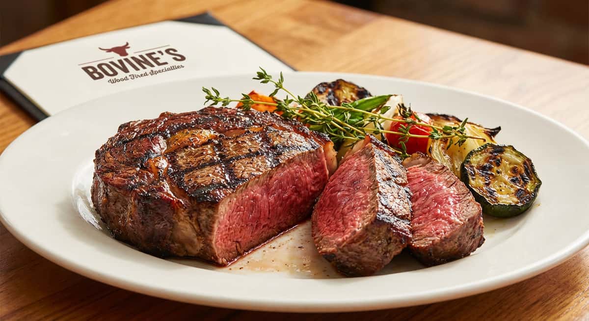 Perfectly grilled ribeye steak cooked medium-rare, with charred vegetables and fresh herbs, served at Bovine's in Murrells Inlet, SC, a premier destination for waterfront dining and wood-fired steaks.