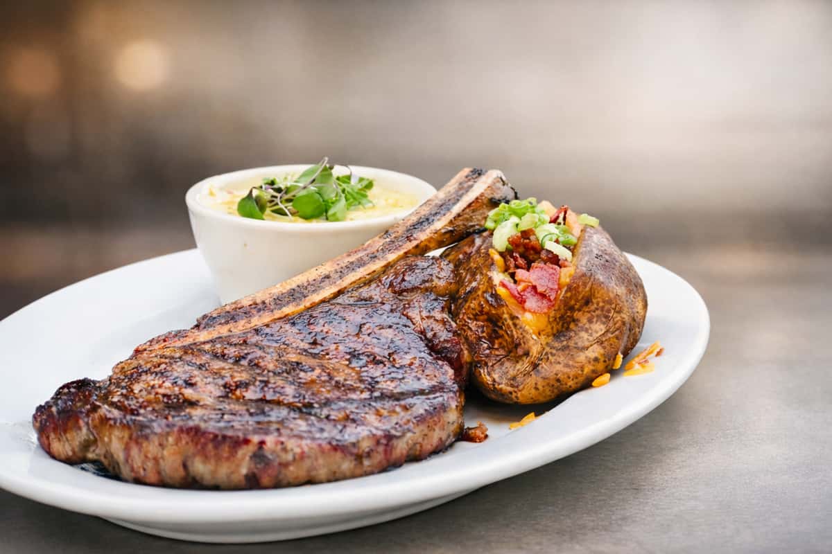 Juicy grilled tomahawk steak served with a loaded baked potato topped with cheese, bacon, and scallions, alongside creamy corn grits garnished with microgreens.