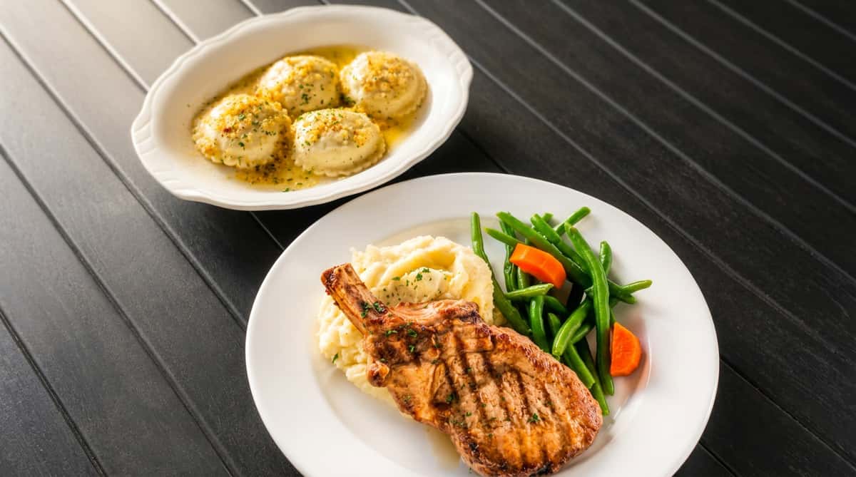 Grilled pork chop with mashed potatoes, green beans, and carrots, alongside ravioli in butter sauce.