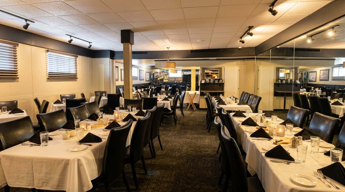 Restaurant dining room with multiple tables set with white tablecloths, black chairs, and dark carpeting.