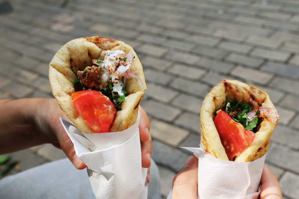 Two hands holding pita wraps filled with fresh tomato, onion, herbs, and a creamy sauce, with a blurred cobblestone background.