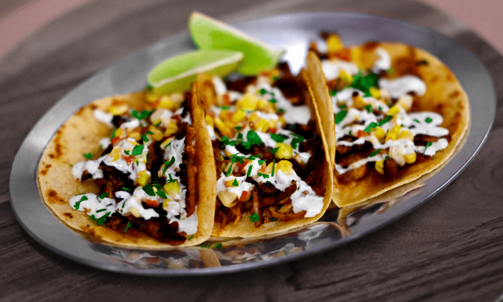 Three street tacos filled with seasoned meat, corn salsa, fresh cilantro, and crema, served with lime wedges on a metal plate.